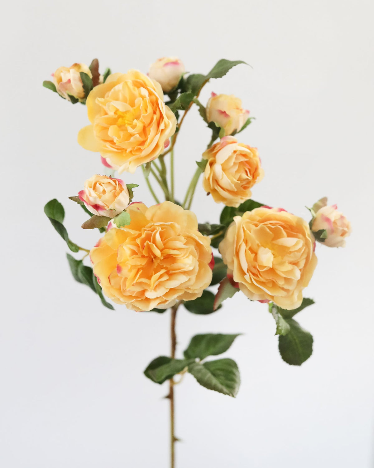 A single stem of artificial yellow-orange roses with green leaves and buds, photographed against a plain light gray background.