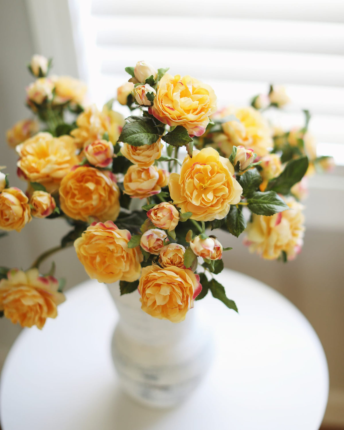 A bouquet of yellow artificial roses with green leaves and pinkish buds, arranged in a white vase on a round white table in front of a softly blurred window.