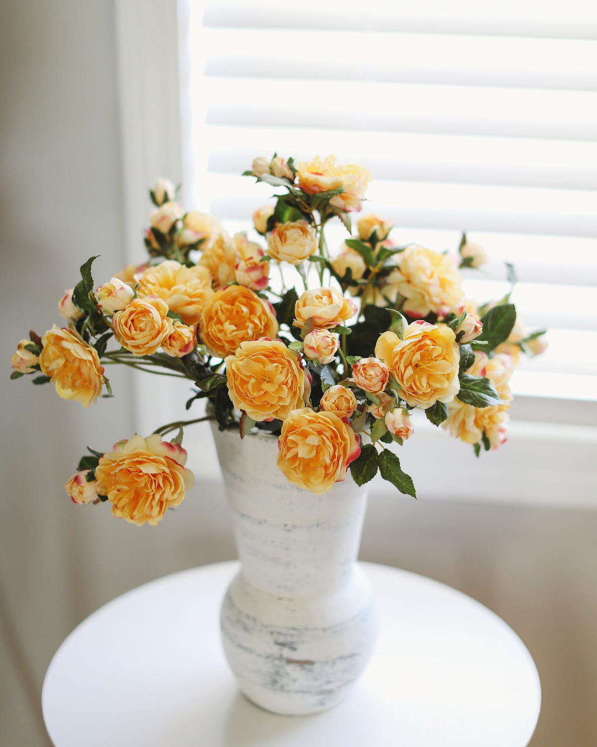 A bouquet of yellow and peach roses with green leaves arranged in a distressed white ceramic vase, set on a white round table in front of a window with white blinds.