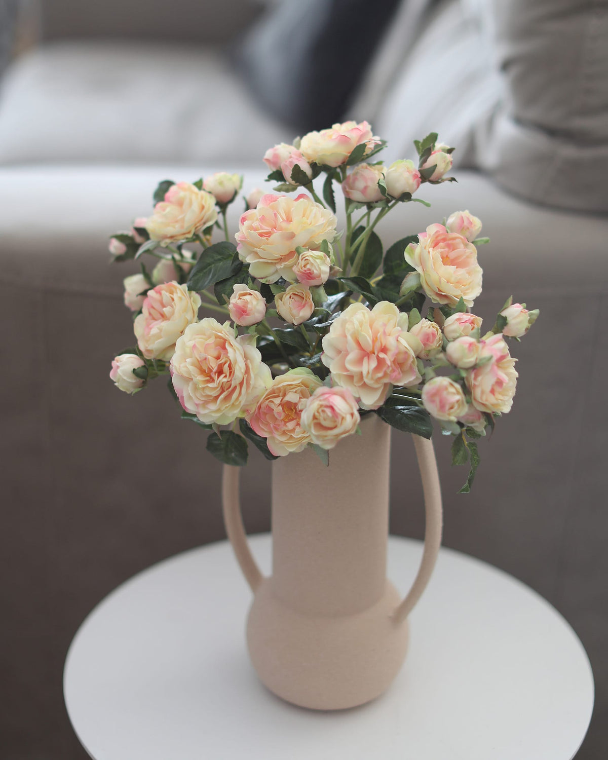 A bouquet of artificial peach and cream roses with green leaves arranged in a beige ceramic vase with side handles, set on a small white round table in a softly blurred living room setting.