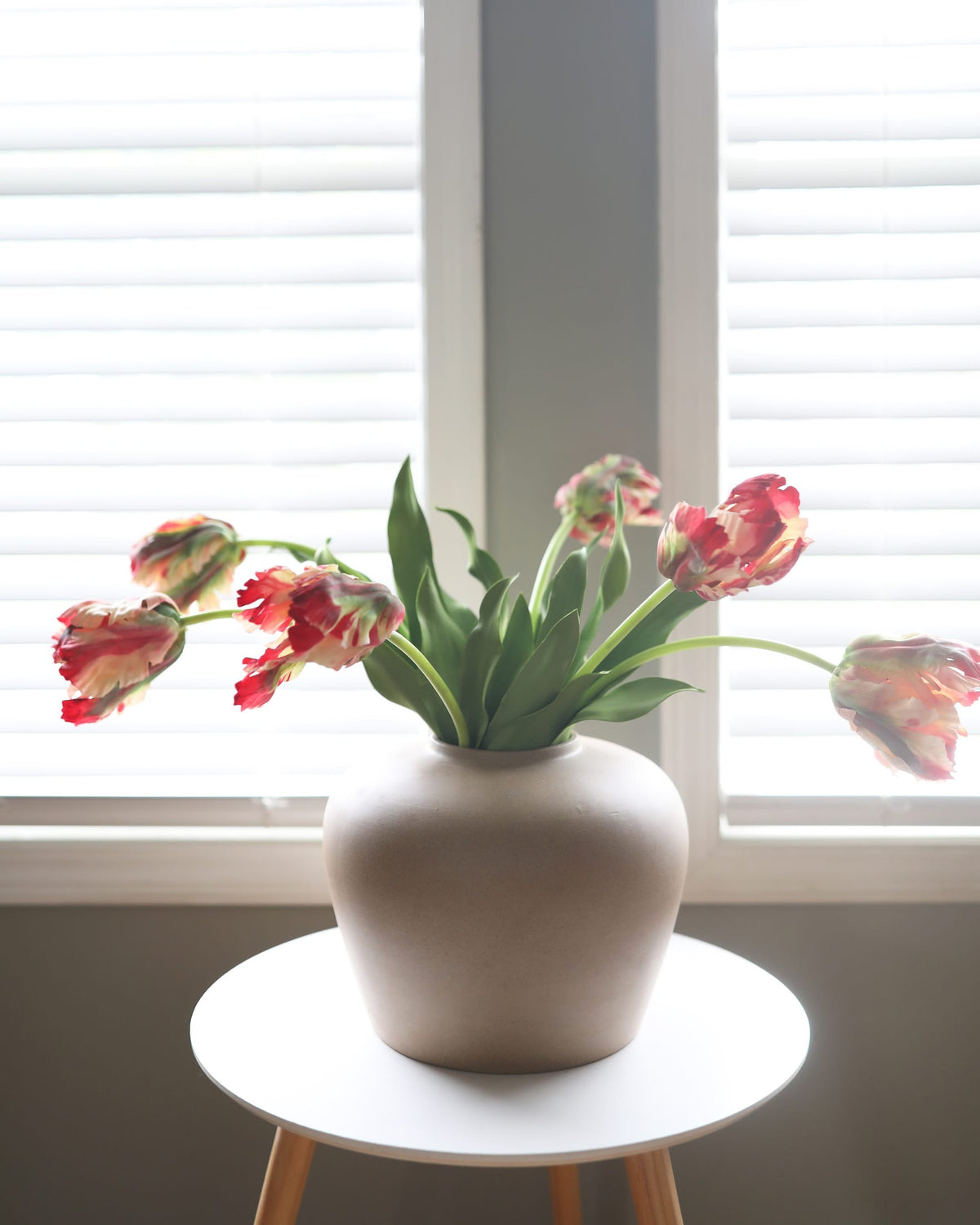 White vase with faux red tulips on a small round table in front of window blinds