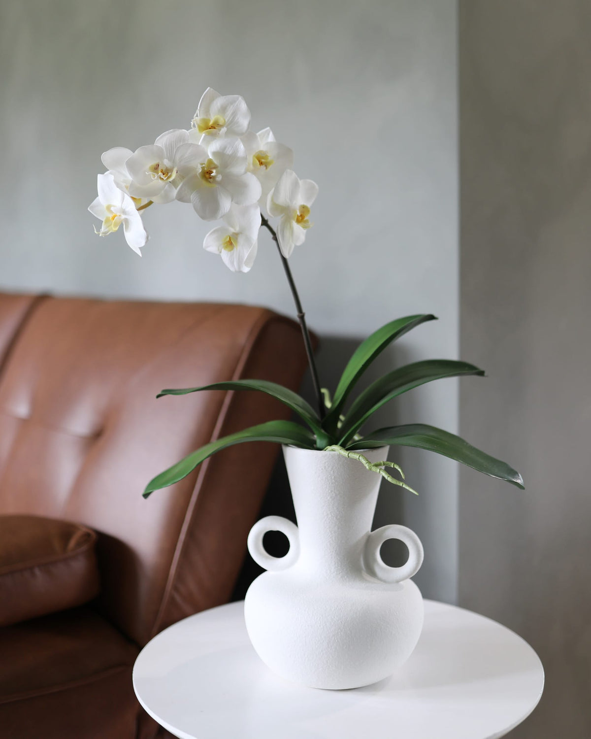 White faux orchid in a modern white vase on a table with a brown leather couch in the background.