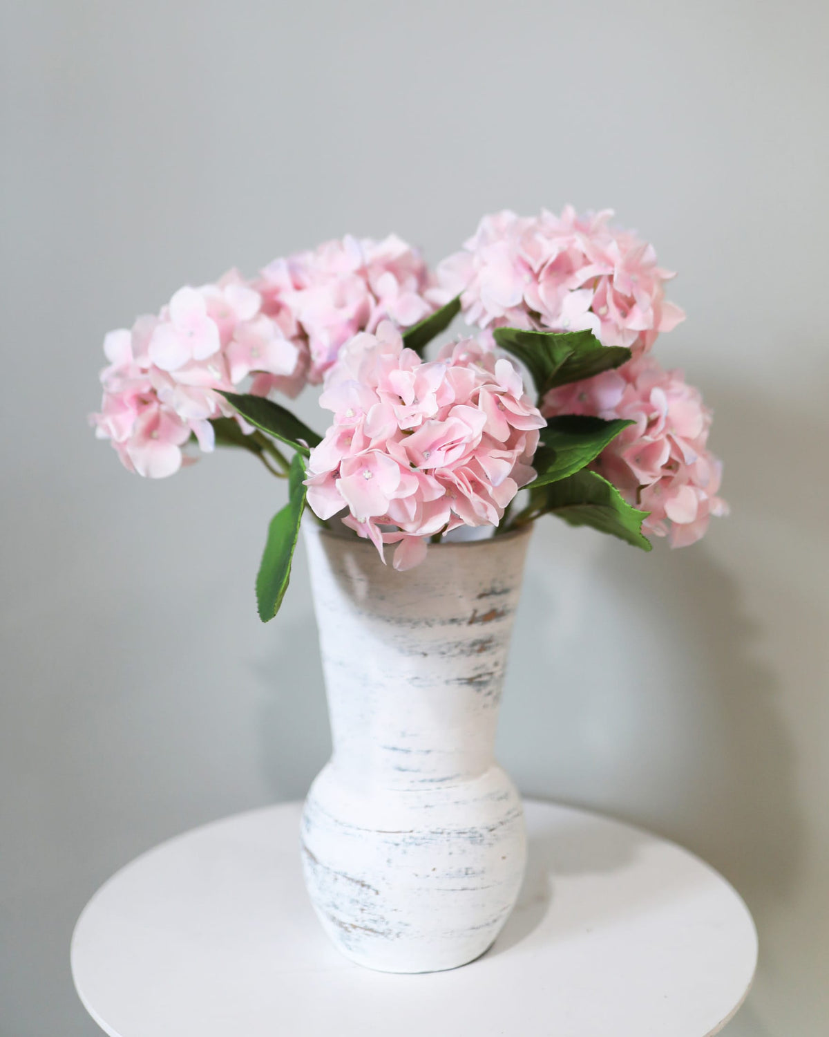 White vase with pink faux hydrangeas on a white surface against a gray background