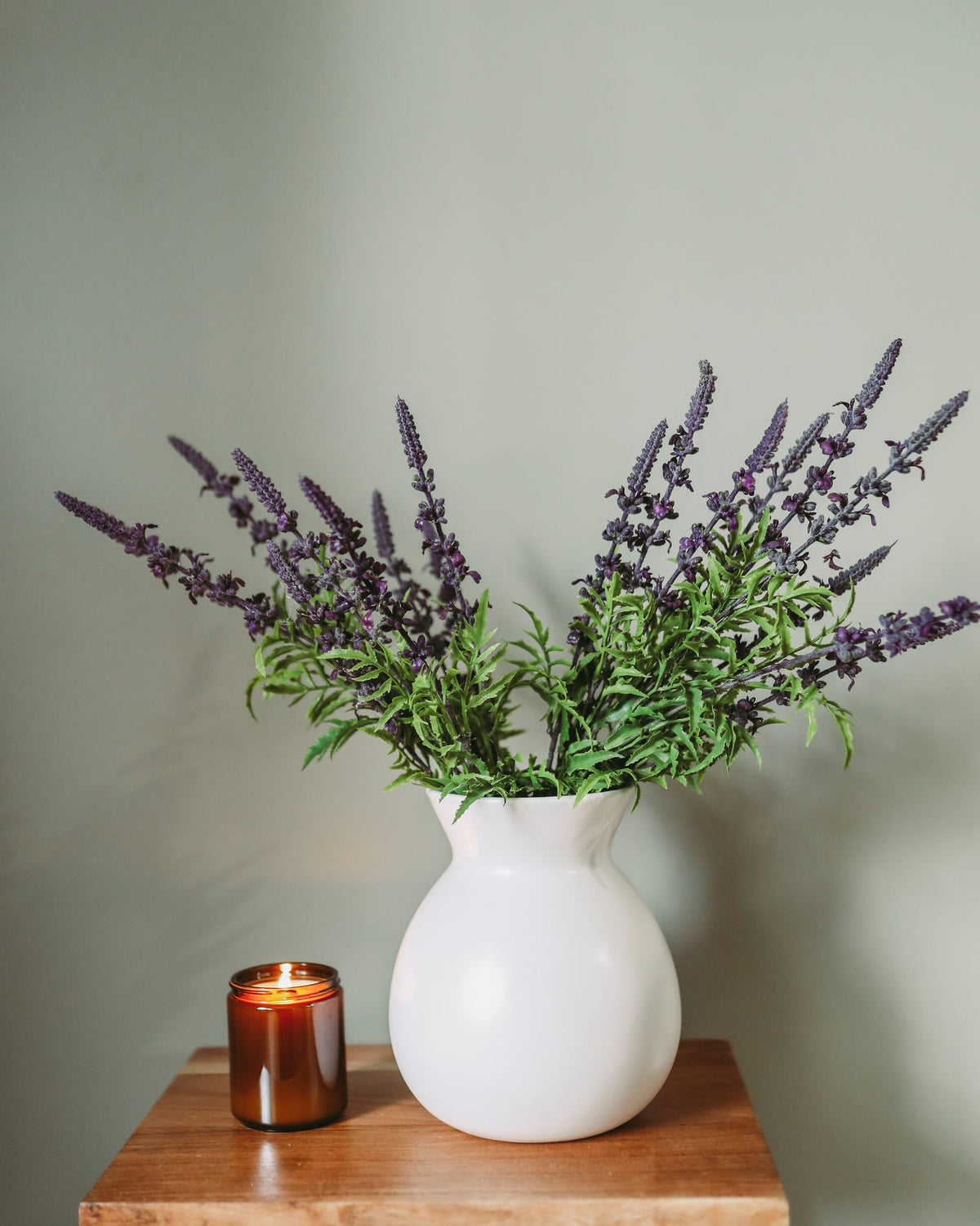 A white ceramic vase filled with purple faux lavender, placed on a wooden table next to a lit amber candle against a muted sage green wall.