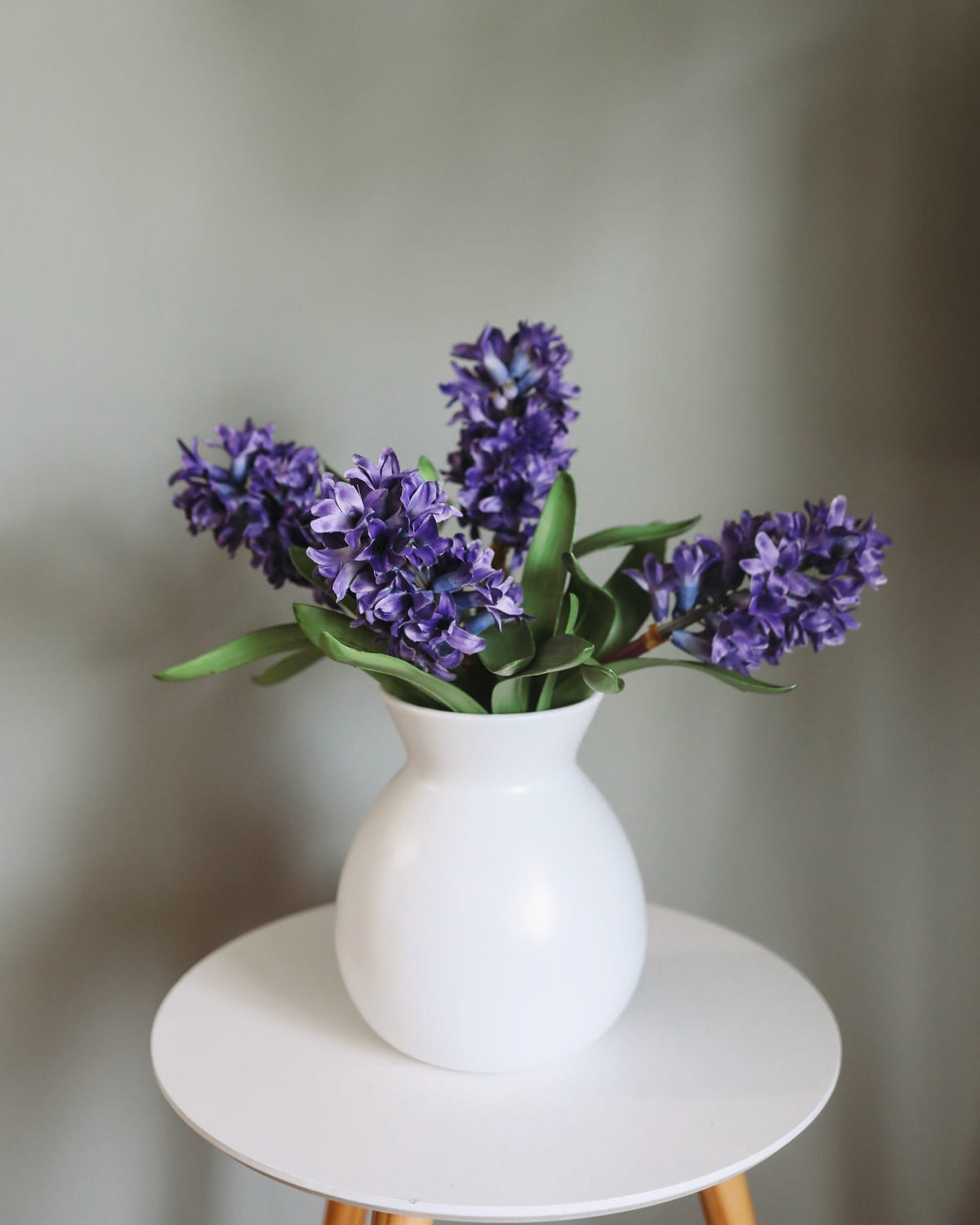 A white ceramic vase filled with faux purple hyacinth flowers, displayed on a small round white side table against a soft gray background.