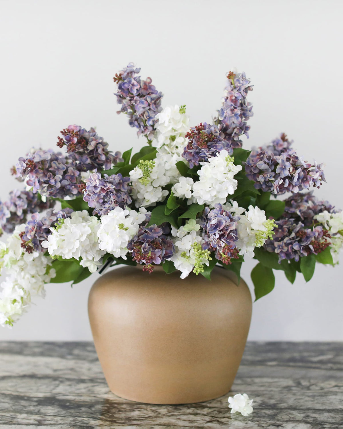 A faux lilac floral arrangement in a matte taupe ceramic vase, set on a rustic gray wooden surface against a plain light gray background.
