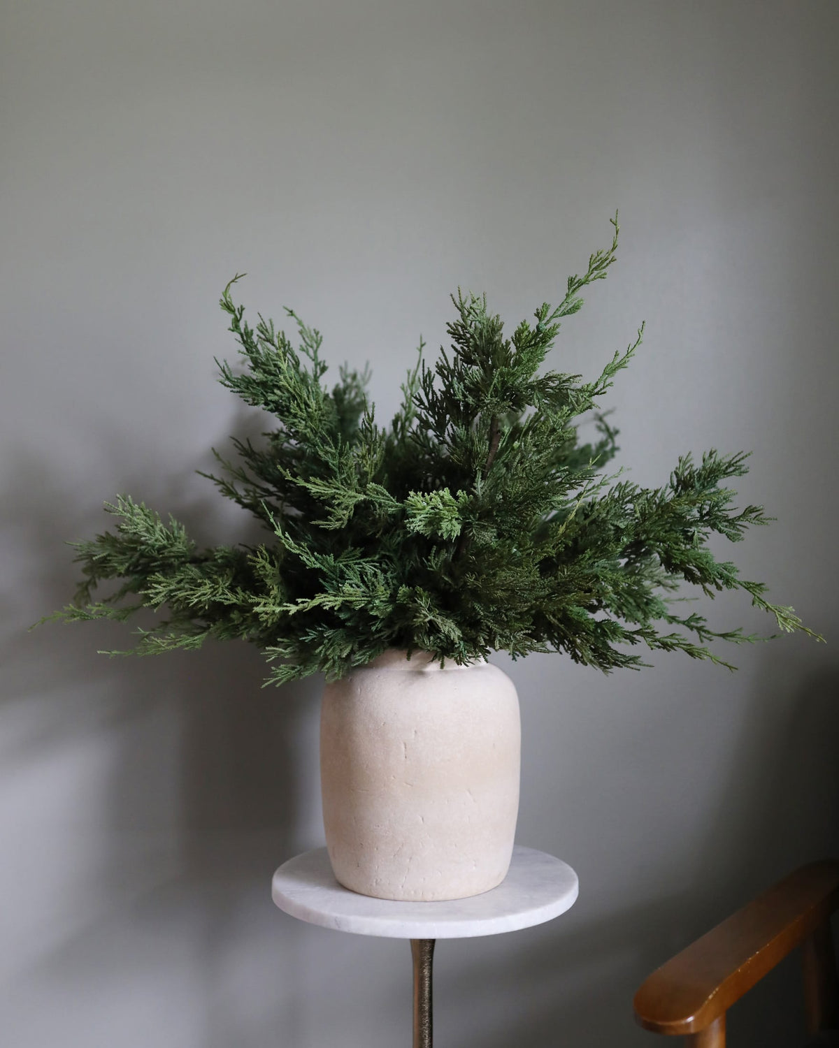 A Faux Cedar Pine branch arrangement in a matte cream ceramic pot, displayed on a small round white side table against a neutral gray wall.