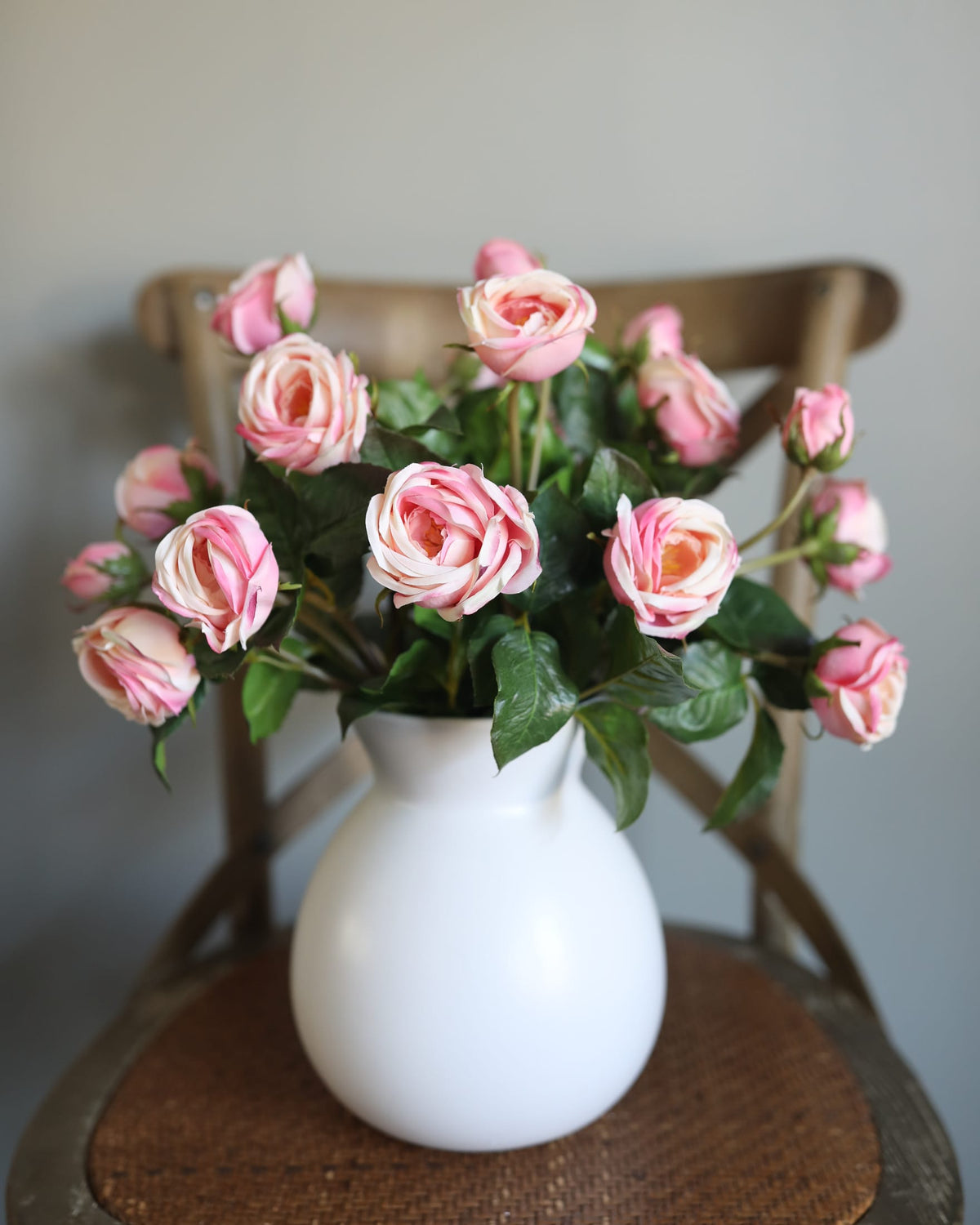 A bouquet of pink and white artificial roses with green leaves arranged in a white ceramic pitcher vase, placed on a wooden chair against a plain gray wall.