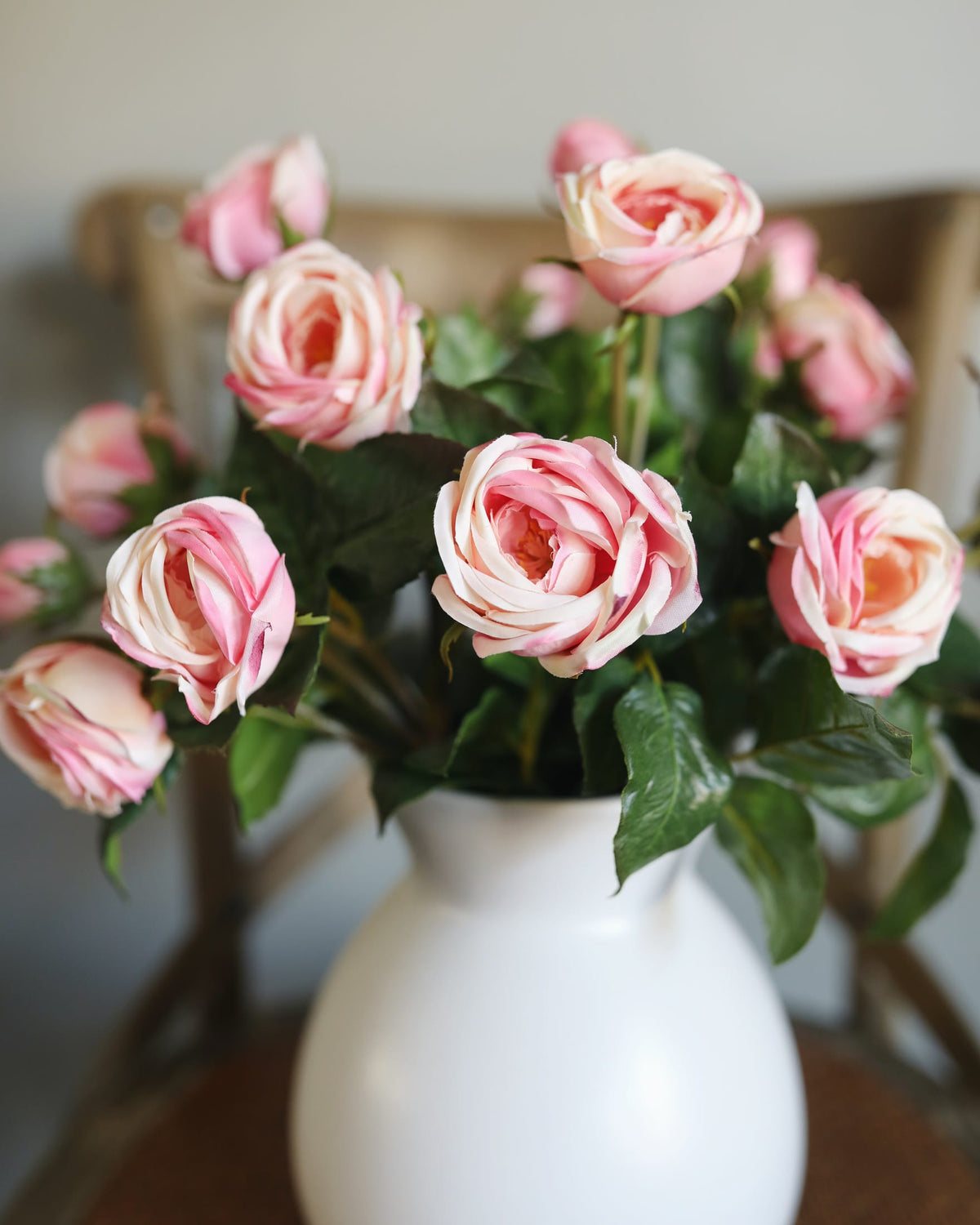 Pink and white striped artificial roses in a white ceramic vase, set against a softly blurred indoor background.