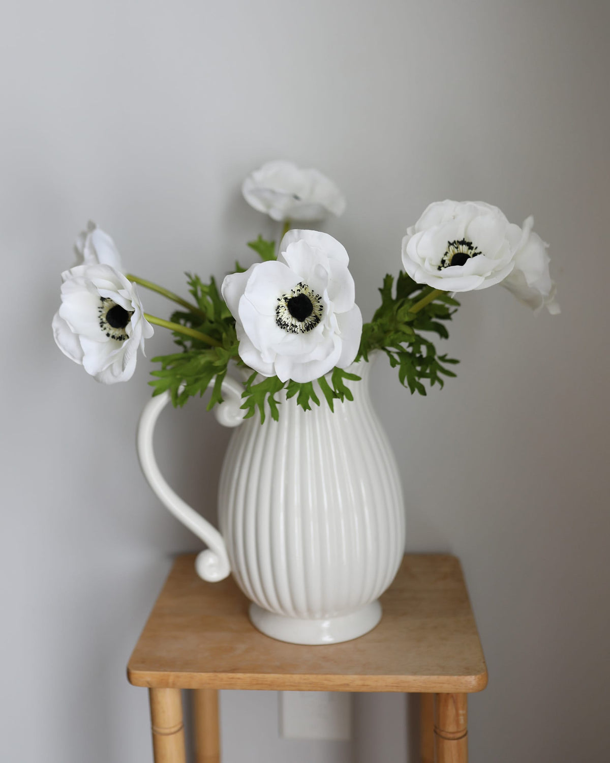 White ribbed ceramic pitcher vase filled with faux white anemone flowers and green leaves, displayed on a small wooden stool against a light gray wall.