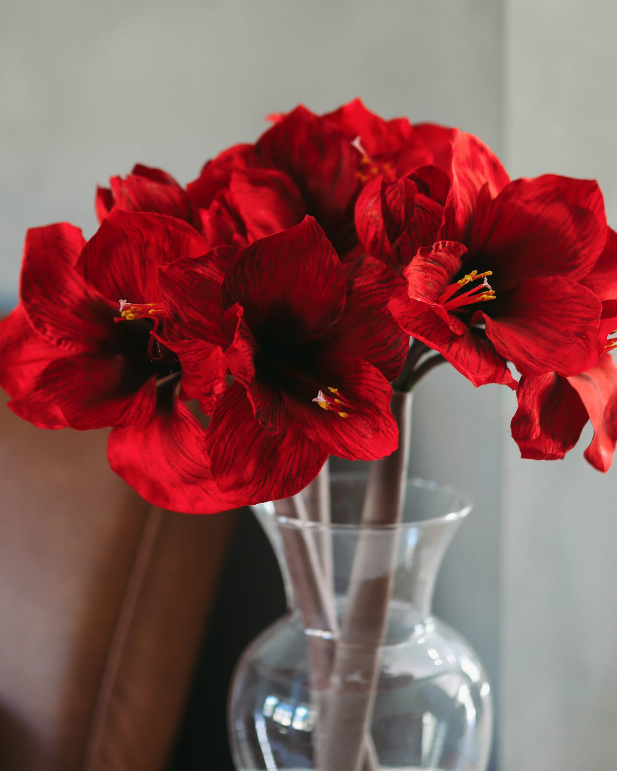 A bouquet of vibrant faux red amaryllis in a clear glass vase, set against a soft-focus background of a gray wall and a brown leather chair.
