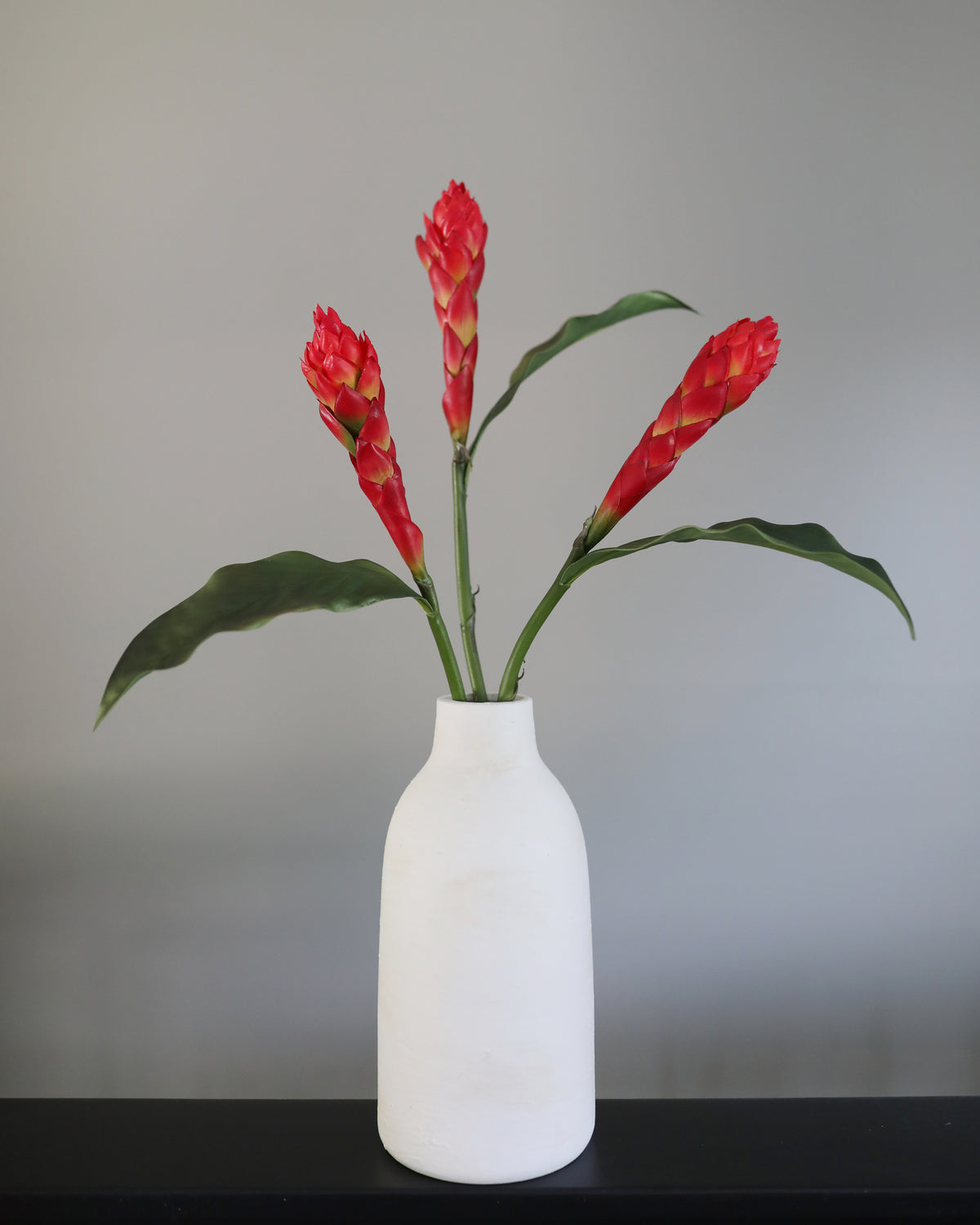 A white ceramic bottle vase holding four bright red-orange tropical gingers with green leaves, set against a plain gray wall and a dark surface.