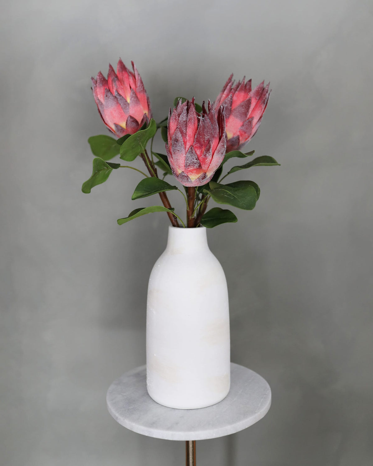 Faux protea flower buds with green leaves in a white vase.