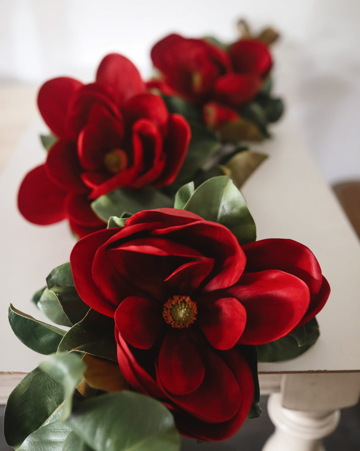 Garland of red magnolia artificial flowers with green leaves arranged on a white table runner in a softly lit indoor setting.