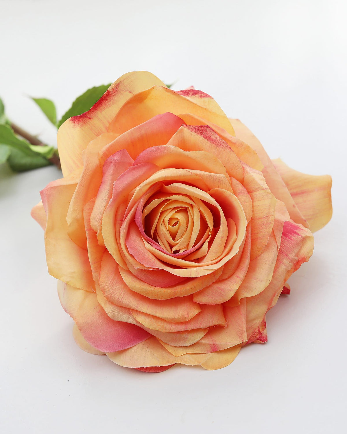 A single artificial rose with peach, orange, and pink petals, shown on a plain white background with a small portion of green stem and leaves visible on the left.