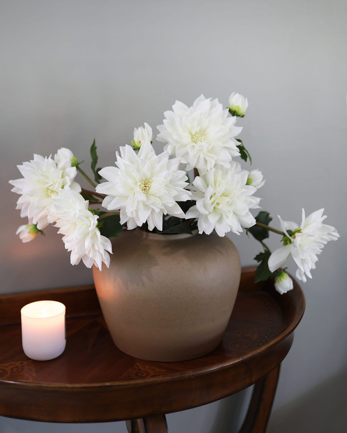 White artificial dahlia flowers arranged in a matte taupe ceramic vase on a dark wood side table, with a lit white candle nearby against a neutral gray wall.