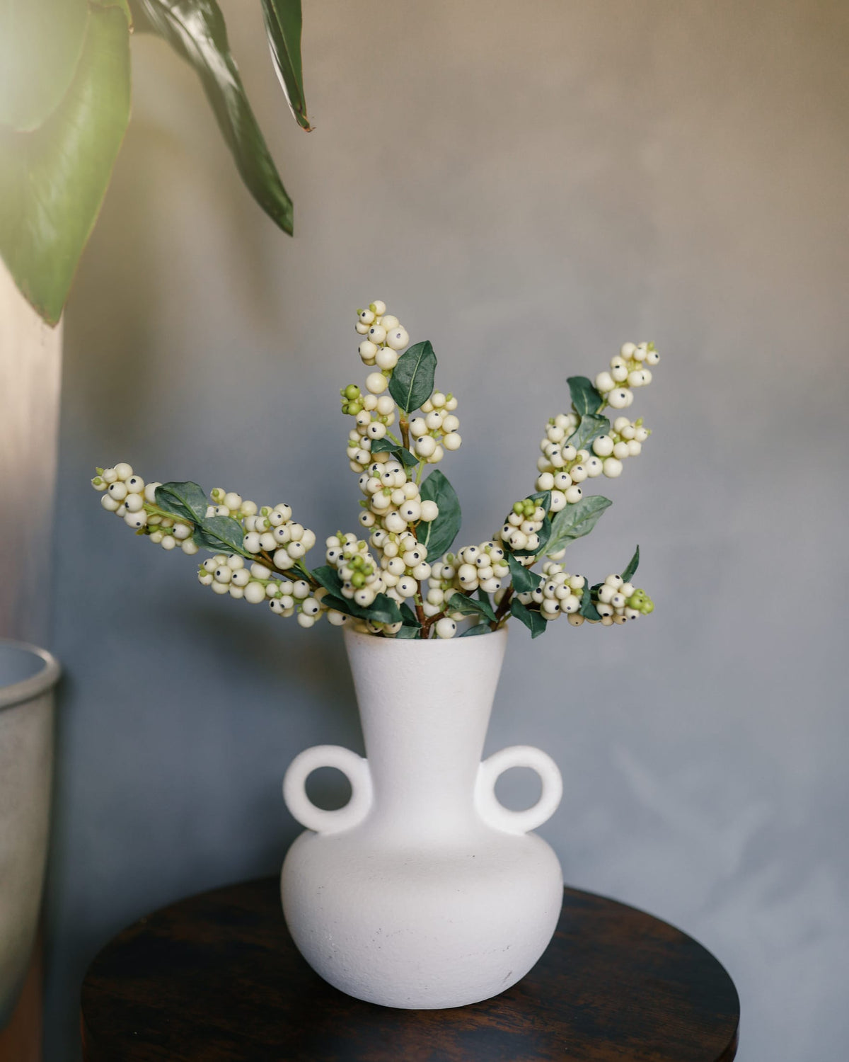 A matte white ceramic vase with two side loops, holding snow berries set on a dark wooden table against a textured gray wall.