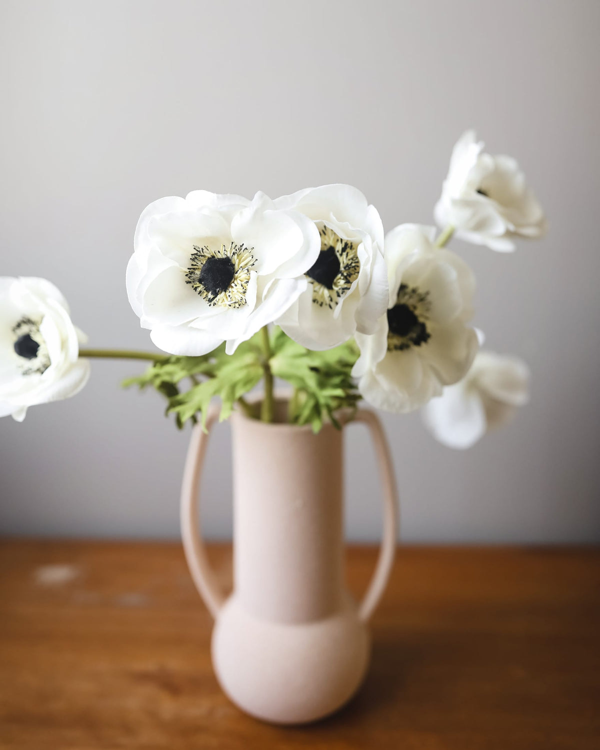 White faux anemone flowers with dark centers arranged in a pale pink ceramic vase with side handles, set on a wooden surface against a soft gray wall.