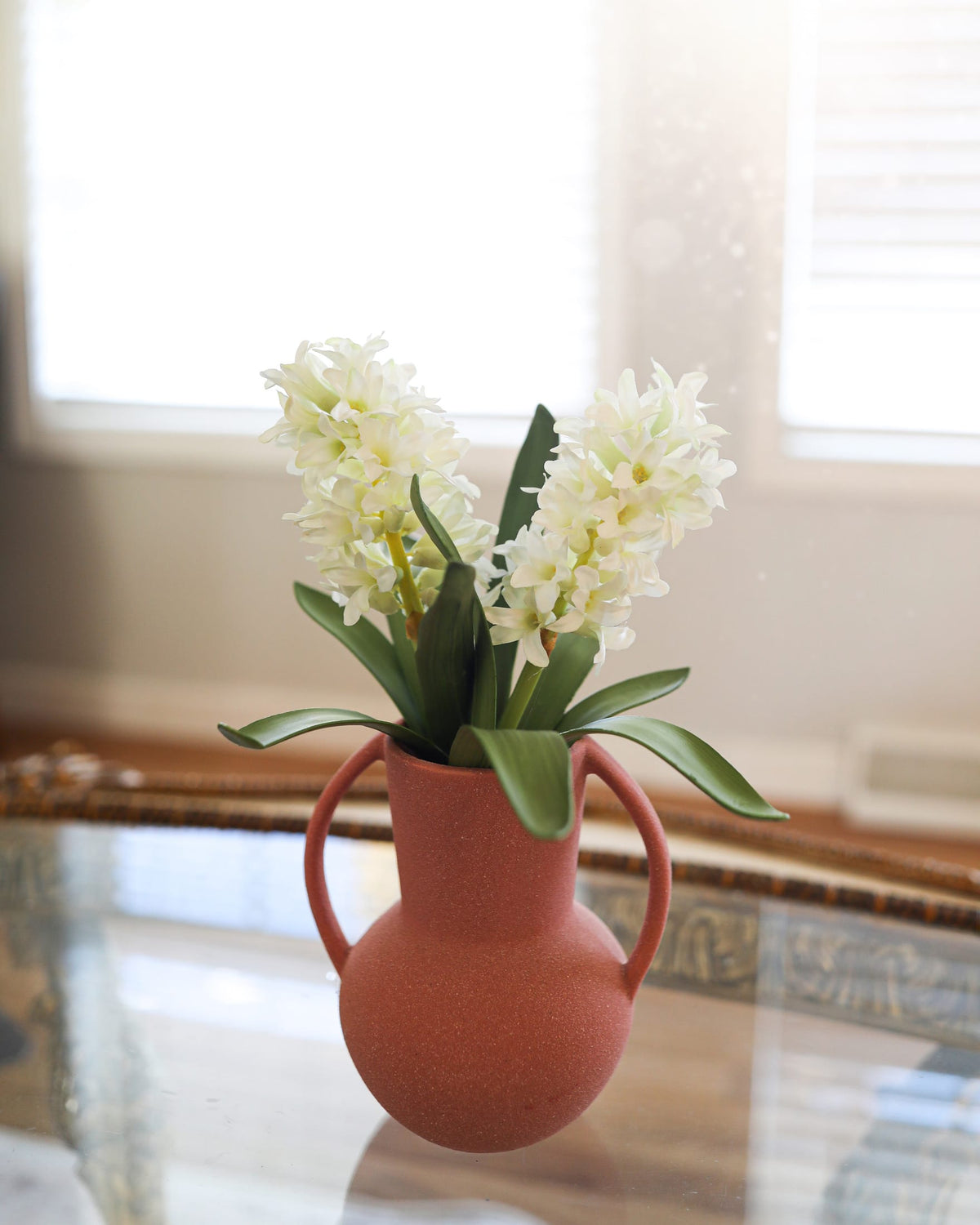 A terracotta-colored ceramic pitcher vase with two handles, filled with faux white hyacinth flowers and green leaves, set on a glass table in a softly lit room.