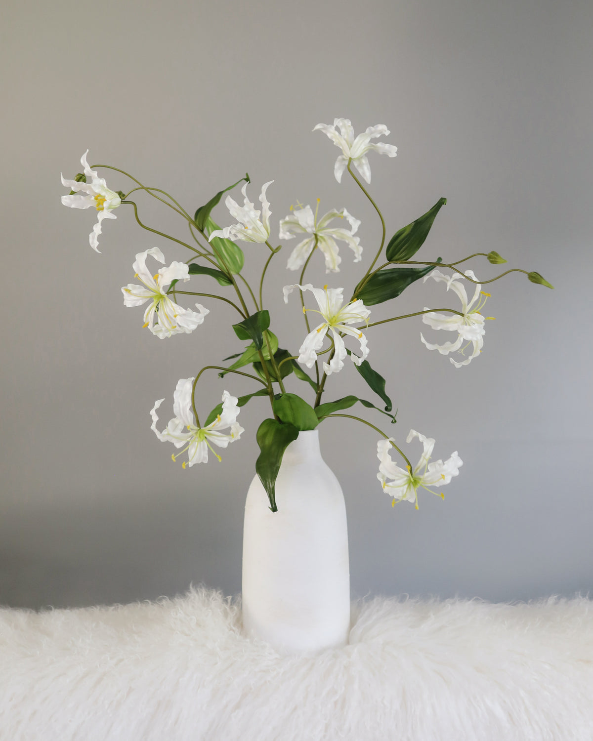 White gloriosa lily flowers with green leaves arranged in a matte white ceramic vase, set on a white faux fur surface against a gray background.