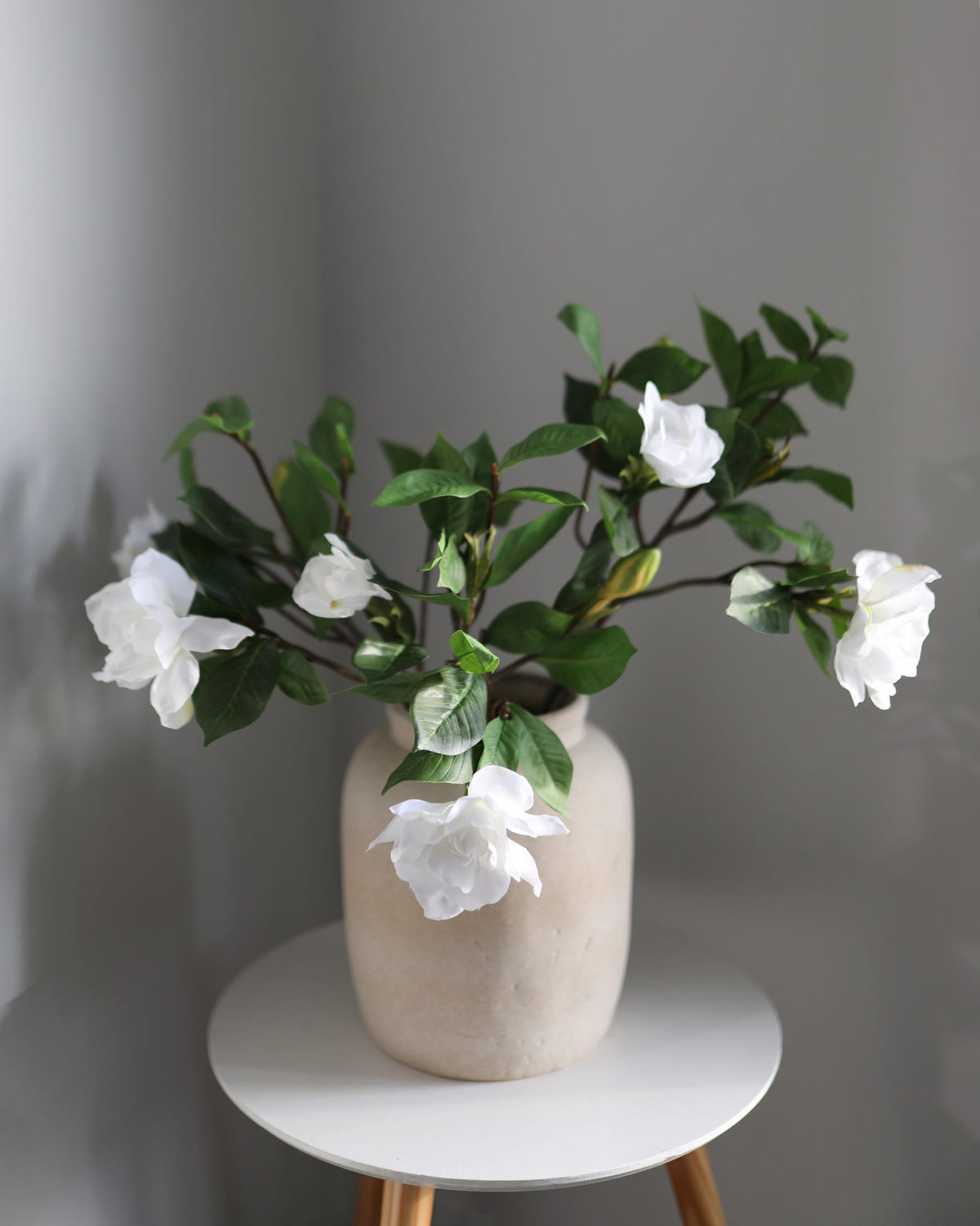 Artificial gardenia in a beige vase on a white stool against a gray wall