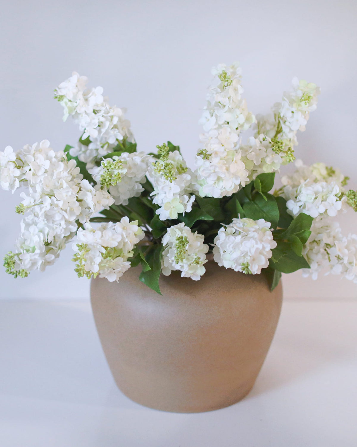 White faux lilac with green leaves arranged in a matte beige ceramic pot against a plain white background.