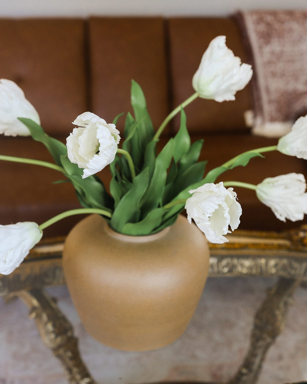 White tulip artificial flowers arranged in a matte beige ceramic vase on an ornate gold table, with a brown leather sofa blurred in the background.