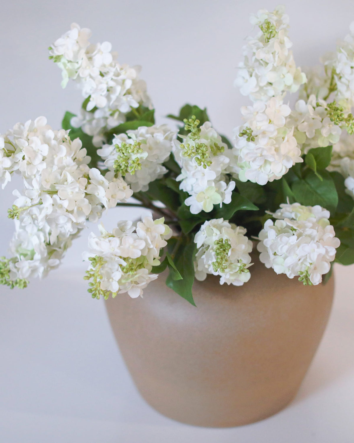 White faux lilac artificial flower arrangement in a matte beige ceramic pot against a plain light gray background.