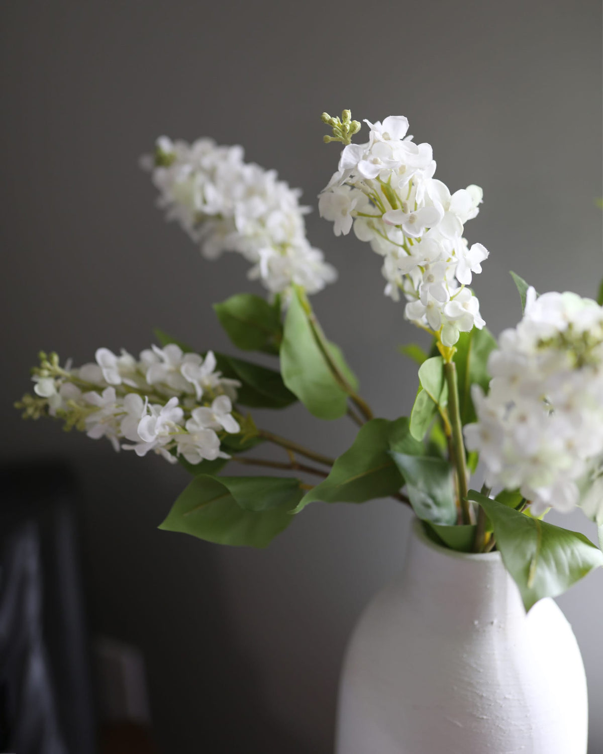 White faux lilac in a white vase against a dark background