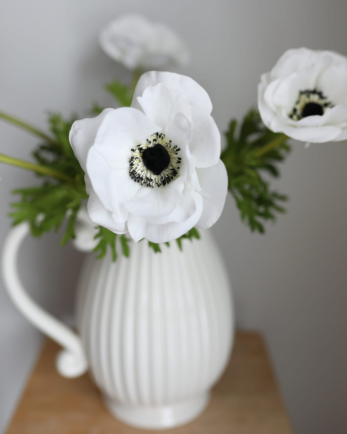 White artificial anemone flowers with dark centers and green foliage arranged in a white ribbed ceramic pitcher on a wooden surface against a neutral gray background.