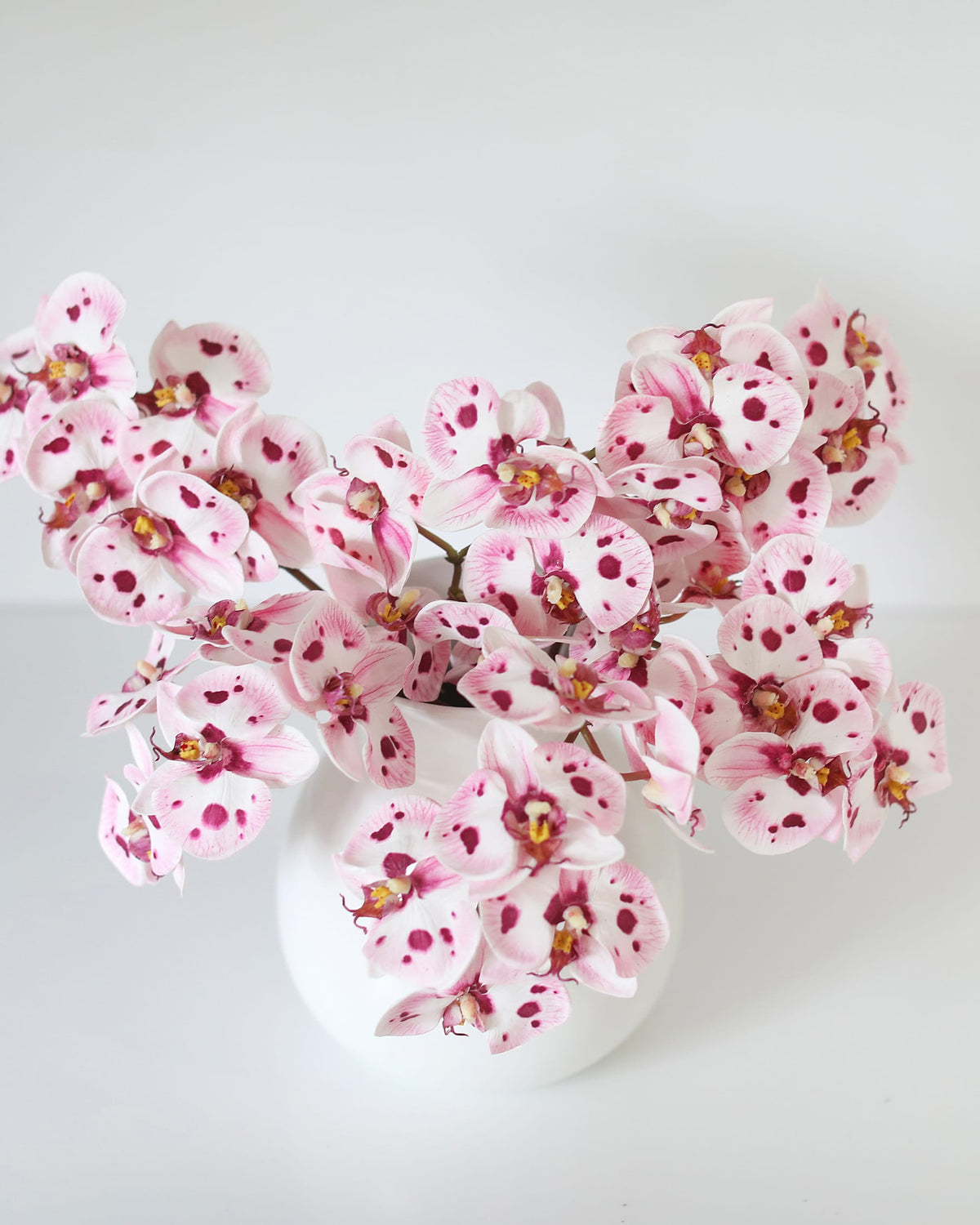 Pink and white faux spotted orchid flowers arranged in a round white ceramic vase against a clean white background.
