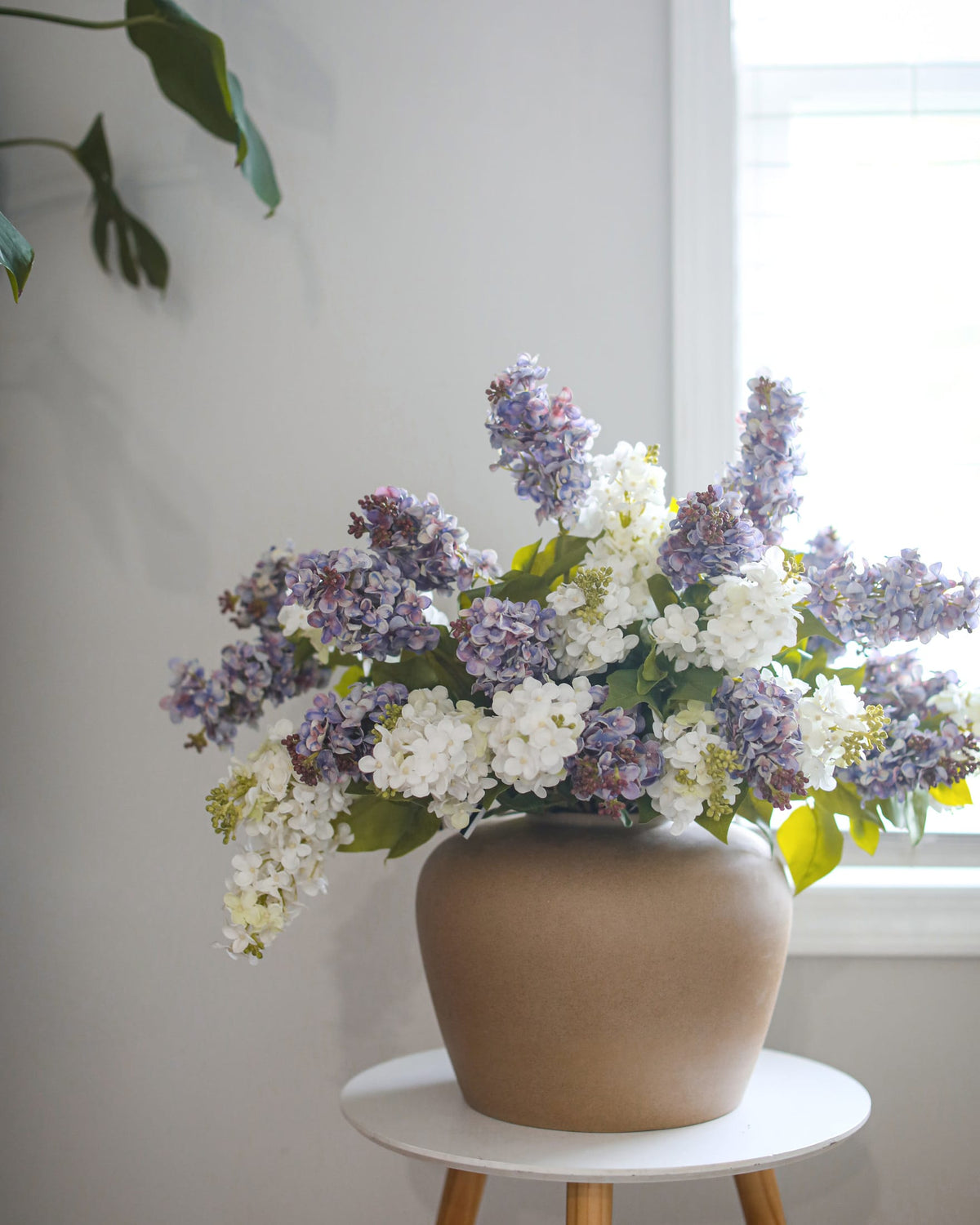 A large ceramic pot in a warm beige tone, filled with a dense arrangement of white and lavender faux lilac with green leaves, set on a small white table in a bright room.