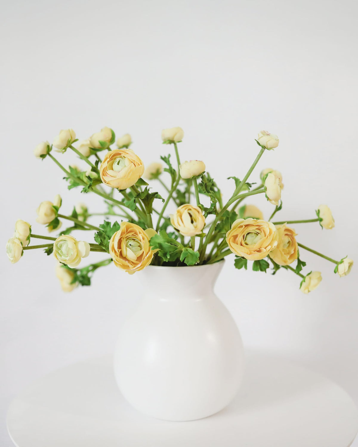 Yellow faux ranunculus flowers with green stems arranged in a matte white ceramic vase, set against a clean white background.